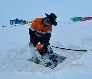 A man wearing high-vis work clothing, earmuffs and a cowboy hat is operating a chainsaw to cut through a thick sheet of ice covering a lake. Behind him, across a stretch of snow-covered ground, are the bright blue, yellow, green and red buildings of an Antarctic station