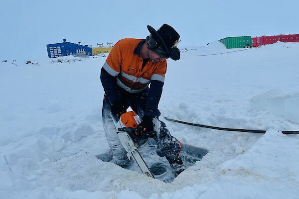 A man wearing high-vis work clothing, earmuffs and a cowboy hat is operating a chainsaw to cut through a thick sheet of ice covering a lake. Behind him, across a stretch of snow-covered ground, are the bright blue, yellow, green and red buildings of an Antarctic station