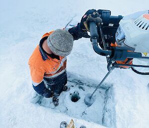 A man in high-vis work clothing and a grey beanie is operating industrial drilling equipment, drilling a hole into the thick ice layer over a lake. The frozen lake surface all around him is covered with snow