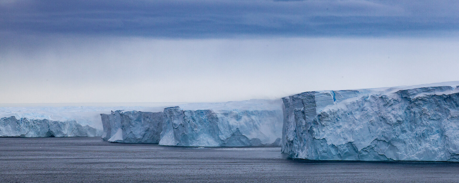 Ice cliffs and ice formations rise out of the water, viewed from a ship.