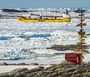 A yellow cargo ship with cranes sits in the water off an icy coastline. A sign in the foreground says "Casey".