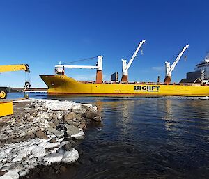 A large yellow ship in a bay prepares to unload cargo using cranes.