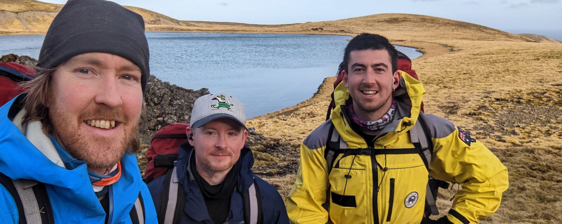 Three men in jackets with back packs stand in front of lake on a grassy peak.