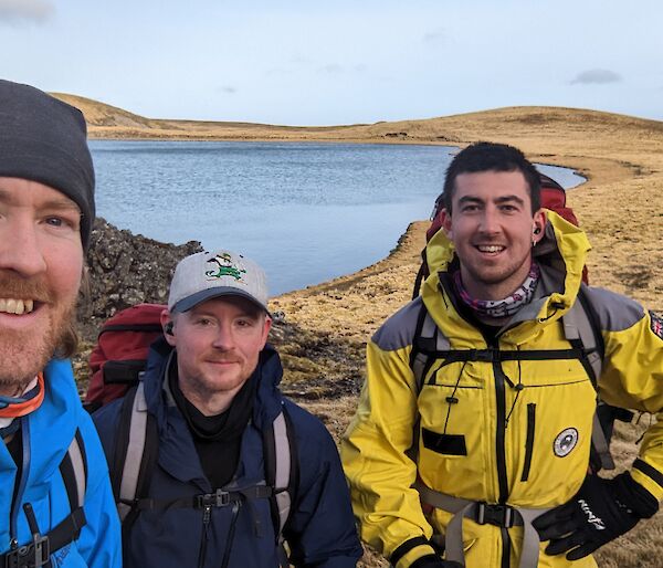 Three men in jackets with back packs stand in front of lake on a grassy peak.