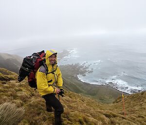 A man in a yellow jacket with a large pack stands on the edge of a grassy hill with the grey ocean making waves on the shore below.