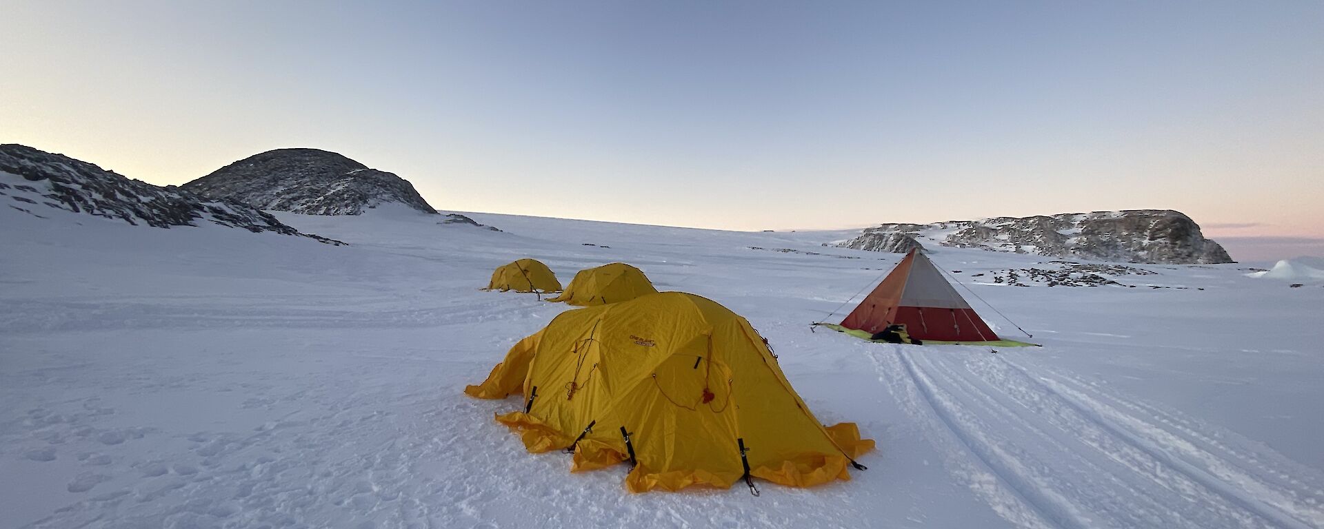 Four tents, three dome shaped and one pyramid, erected on area of flat snow covered ground. In the distance sunset brings a faint orange glow to the horizon.