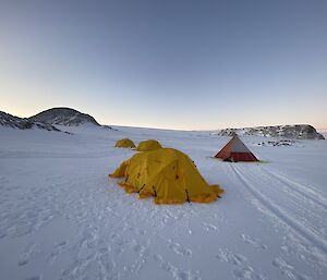 Four tents, three dome shaped and one pyramid, erected on area of flat snow covered ground. In the distance sunset brings a faint orange glow to the horizon.