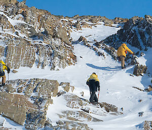 Three people wearing yellow jackets climb up rocky snow covered slope