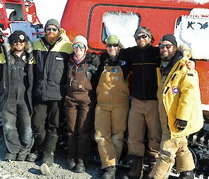 Group of six expeditioners standing in front of Hägglunds, with arms around shoulders and smiling towards camera.