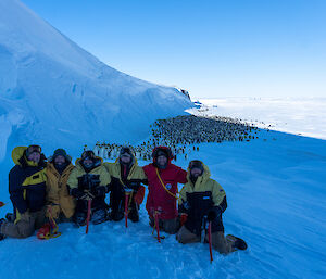 Group of six expeditioners in foreground, in the distance an emperor penguin colony pushed up against ice cliffs.