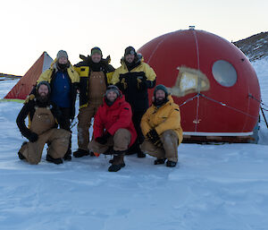 Six expeditioners pose in front of a round red field hut