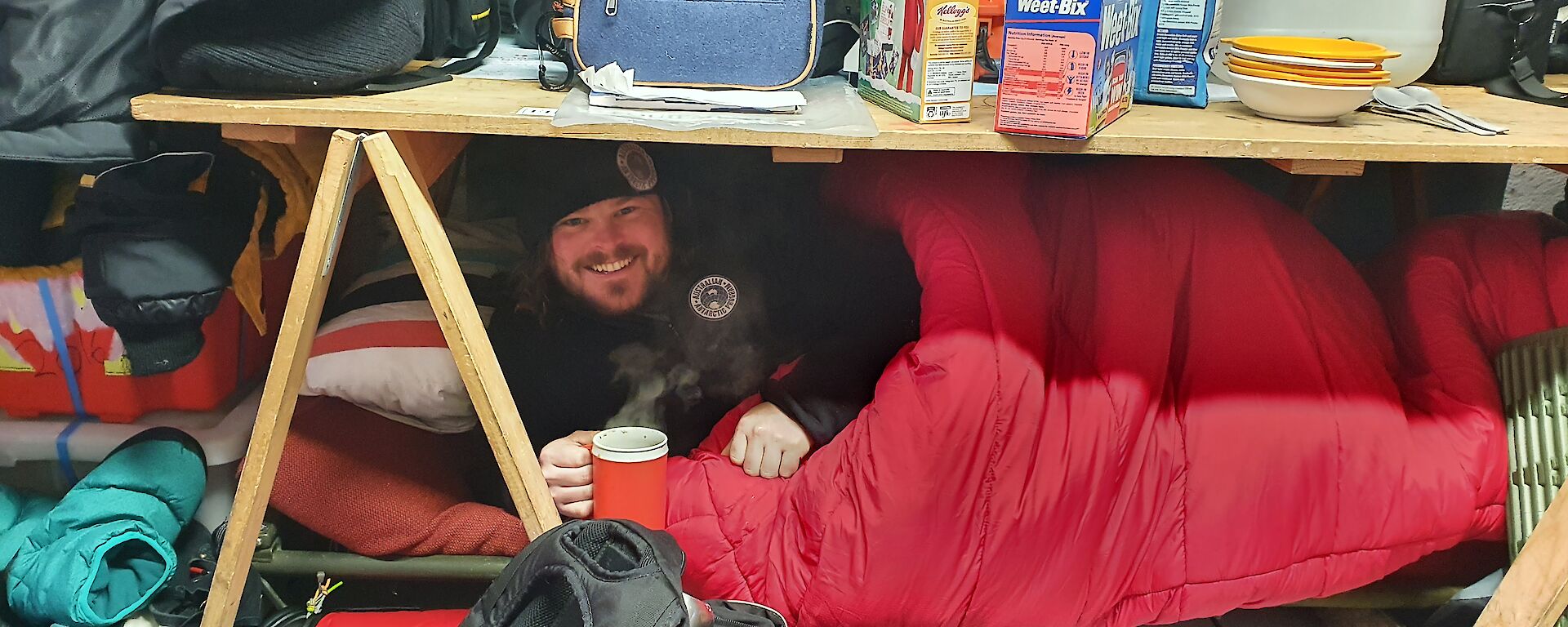 Man in sleeping bag smiles to camera, holds cup of steaming liquid, whilst under a wooden trestle table.