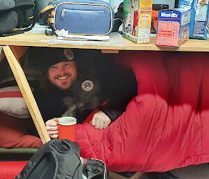 Man in sleeping bag smiles to camera, holds cup of steaming liquid, whilst under a wooden trestle table.