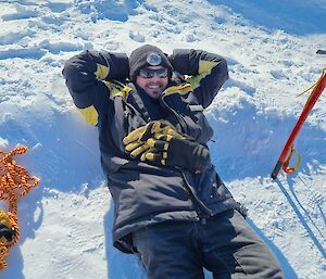 A man with sunnies and a beanie lies on his back in the snow smiling up a camera