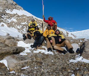 Six expeditioners group around marker point on rocky outcrop with patches of snow