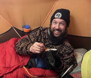 Man lies in sleeping bag in orange tent, smiles to camera, and holds an open bag of dehydrated food.