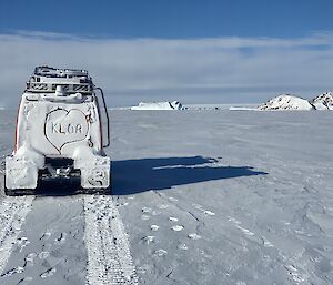 In foreground rear of Hägglunds vehicle with KLOA written into the snow on the back window inside a love heart. Hägglunds travels across flat sea ice with icebergs and rocky island in distance.