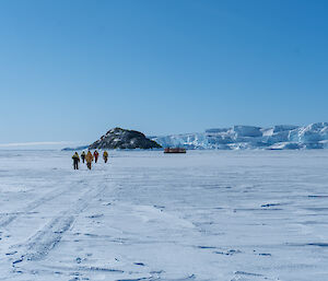 Group of expeditioners walking in the distance away from camera towards Hägglunds vehicles which are parked in front of rocky island and ice cliffs.