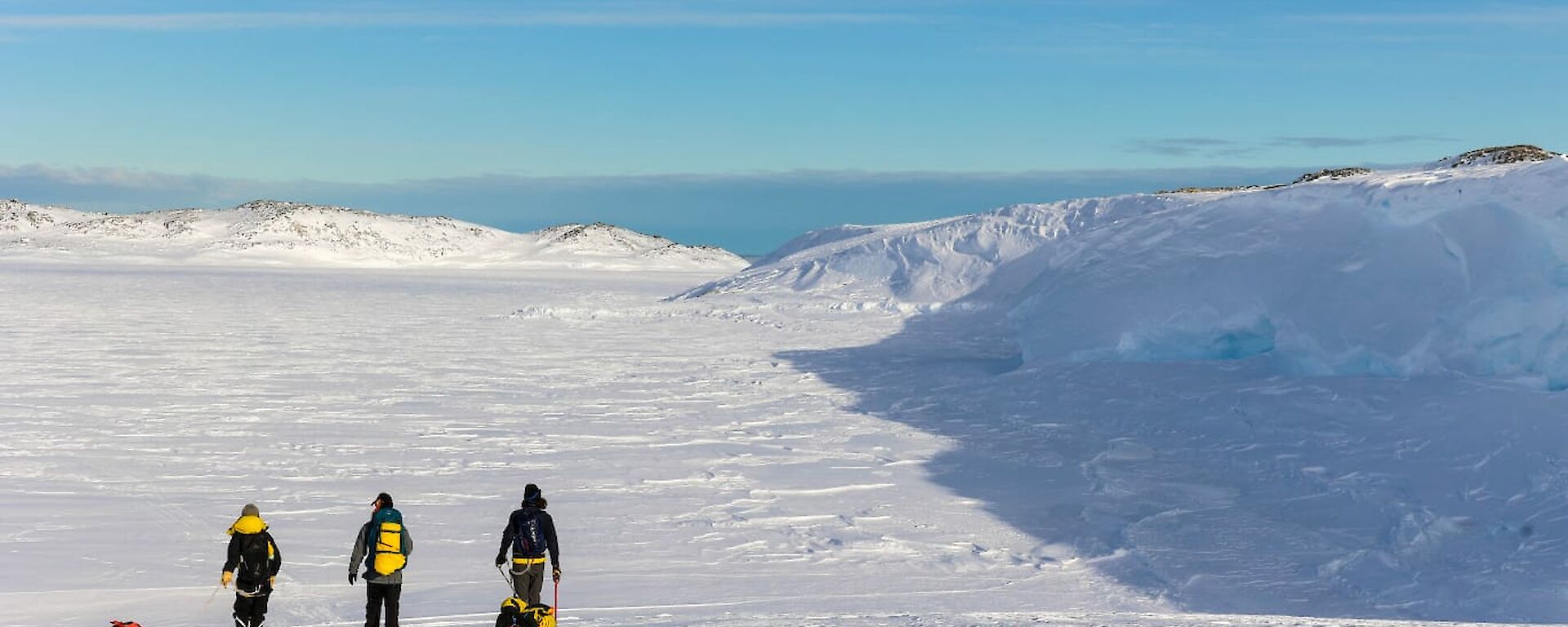 A view from behind of three people walking across a snowy plain bordered with low hills. Two of the people are pulling sleds loaded with gear, the third carries a large pack on his back. The sky is bright blue fringed with grey clouds.