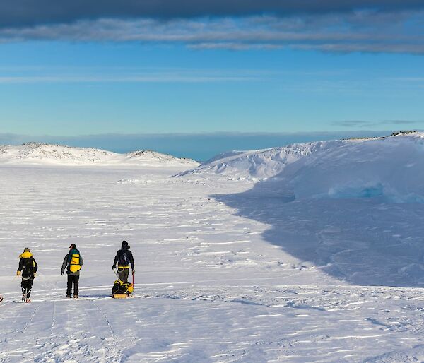 A view from behind of three people walking across a snowy plain bordered with low hills. Two of the people are pulling sleds loaded with gear, the third carries a large pack on his back. The sky is bright blue fringed with grey clouds.