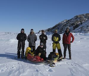Six people posing for a group photo before a snow-dusted hill of jagged, black rock. One person crouches in front beside a couple of sleds loaded with gear, while the other five stand behind her. The sky is bright, clear blue.