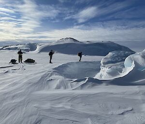 Three people walking across snow-covered ice beneath soft blurs and streaks of high cloud. The snow has been carved by the wind into ridges and scours curving around trapped icebergs.