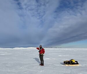 A man in a bright red jacket with a camera hanging around his neck poses for a photo. He is standing on a wide, snowy plain and is pulling a sled loaded with gear on a rope tied around his waist. Soft wings of cloud seem to radiate from the horizon behind him.