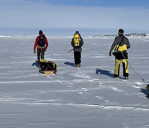 A view from behind of three people walking across a snowy plain. Two of the people are pulling sleds loaded with gear, the third carries a large pack on his back.
