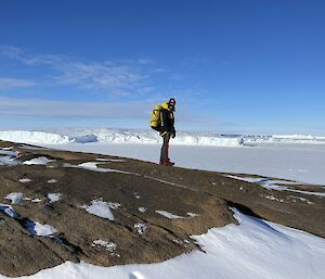 A man wearing a large pack and carrying an ice axe stands on sloping rock above a frozen white sea. In the background is a line of icy coastal cliffs, marbled blue and white.