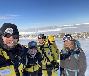 Four people wearing packs and thick winter clothing, posing for a photo on the sea ice on a bright, sunny day.