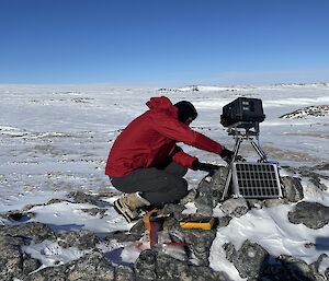 A man in a red weatherproof jacket crouches as he sets up a rugged camera, mounted on a tripod and powered by a small solar panel. The camera stands on a rocky rise above a snowy plain.