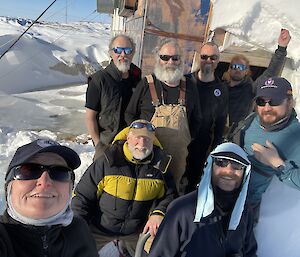 Eight people gathered for a photo in front of a weather-beaten wooden hut with a huge amount of snow banked against it.