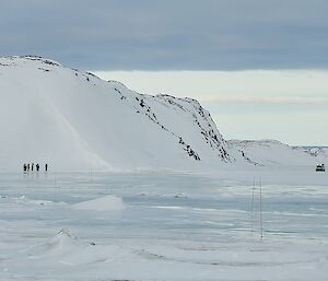 The sea ice is beginning to melt and water is visible on the top. A Hägglunds and a group of people can be seen in the distance.