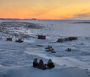 Three people sitting on snow looking at the sunset over a series of machines and containers parked on the snow