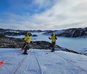 Two people standing in yellow jackets are connected to ropes on the top of an ice cliff