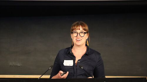 A woman with a fringe and glasses stands at a lectern delivering an address