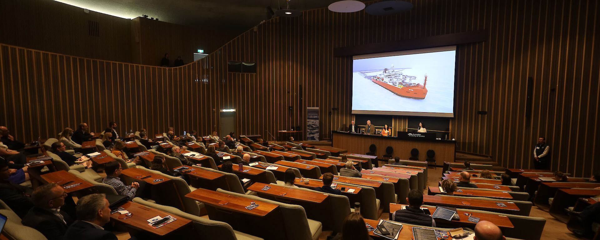 A auditorium filled with people seated. A picture of Australia's icebreaker RSV Nuyina is on the projector screen.