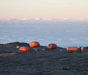 A series of round, orange huts on a rocky coastline with sea ice behind