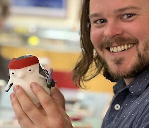 Close up photo of man smiling at camera holding up plastic penguin in left hand and paintbrush in right