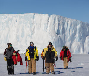 Four expeditioners walk towards camera; dwarfed by a large tabular ice berg in the distance