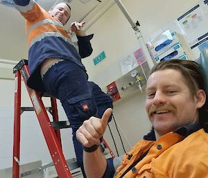 Two men in work clothing, one of them standing on a ladder, making surfers' shaka hand gestures and smiling at the camera