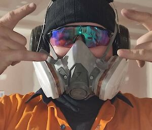 A man in an orange work shirt, protective eyewear, earmuffs, and a respirator mask , making "rock on" gestures with both hands at the camera