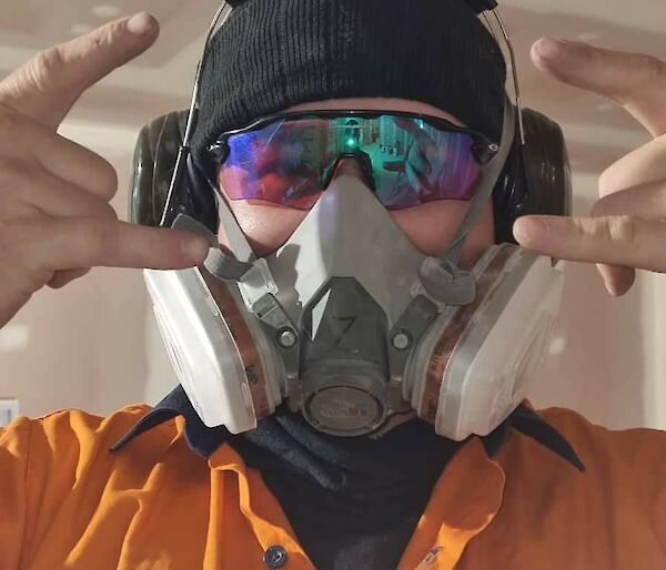 A man in an orange work shirt, protective eyewear, earmuffs, and a respirator mask , making "rock on" gestures with both hands at the camera