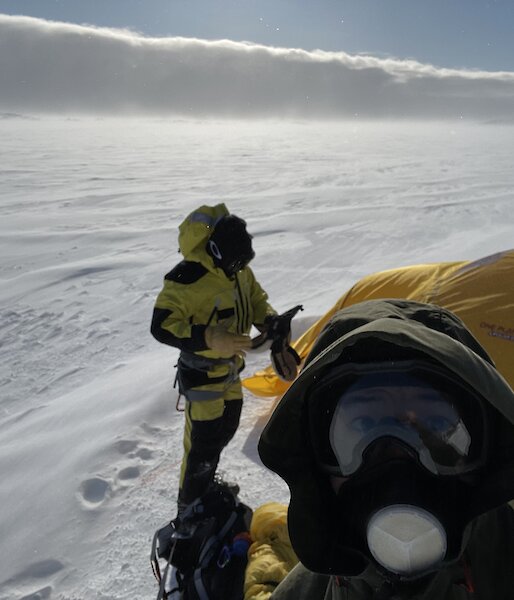A selfie of two men in outdoor hiking gear with a yellow tent on the snow