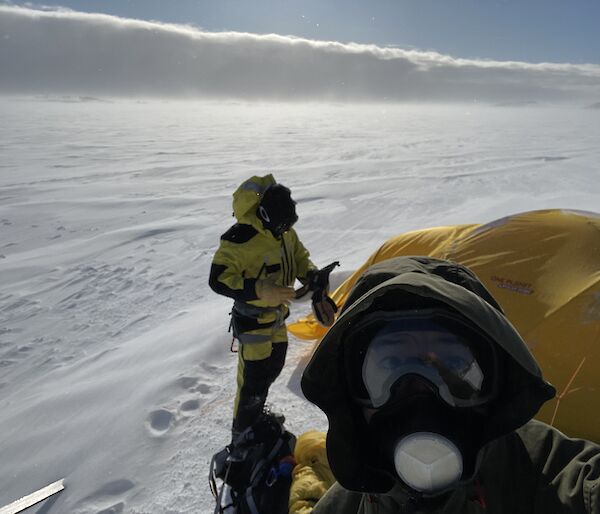 A selfie of two men in outdoor hiking gear with a yellow tent on the snow