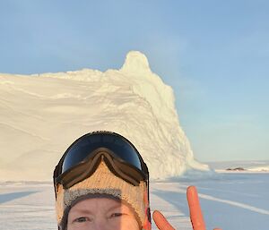 Woman standing in front of iceberg smiling and raising two fingers as victory sign