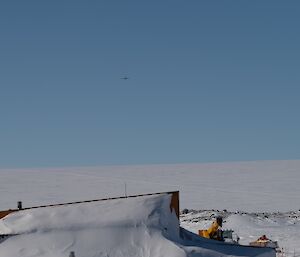 A plane high in the sky above the distant horizon, seen from amidst the snow-covered buildings of an Antarctic station