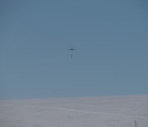 A cargo plane high in a pure blue sky above a snowy plain. Two parcels with parachutes attached have just been ejected from the plane