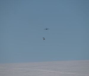 A cargo plane high in a pure blue sky above a snowy plain. Two parcels just ejected from the plane are floating to the ground on parachutes