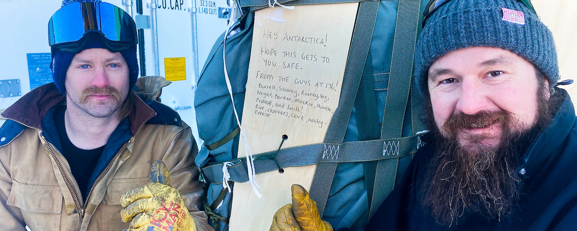 Two men crouching beside a large, cloth-covered parcel bound with several straps, giving thumbs-up gestures to the camera. A plank of wood strapped against the parcel bears a handwritten message that reads: "Hey Antarctica! Hope this gets to you safe. From the guys at 176!" The message is followed by a list of names